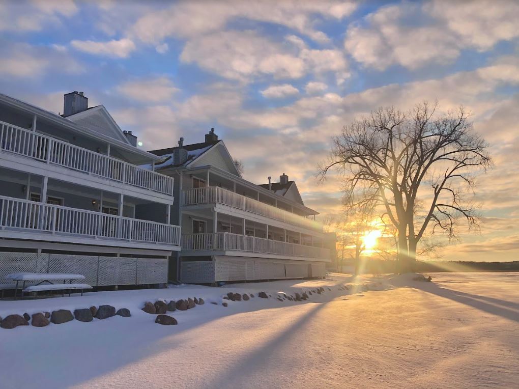 The sun is setting behind a snow covered building with a tree in the foreground.