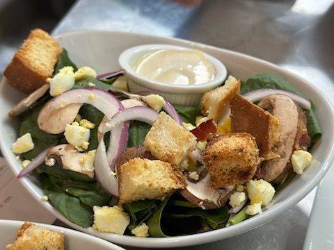 A salad with croutons , eggs , spinach , mushrooms and onions in a bowl on a table.