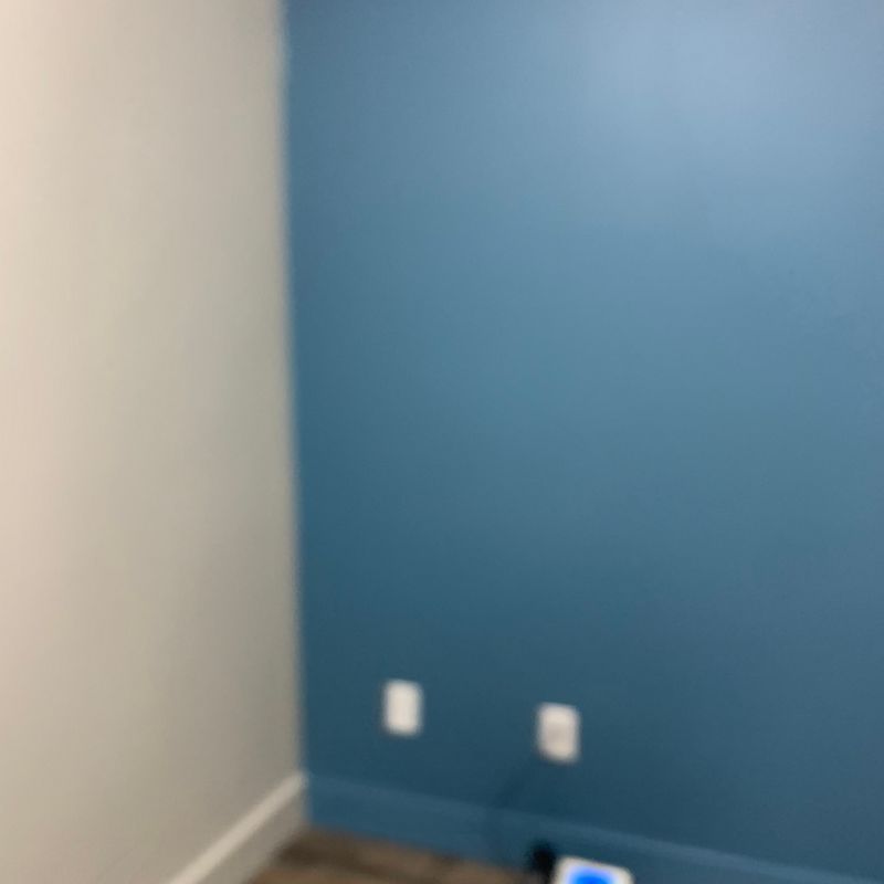 Empty room corner with blue accent wall, beige wall, two outlets, and baseboard.