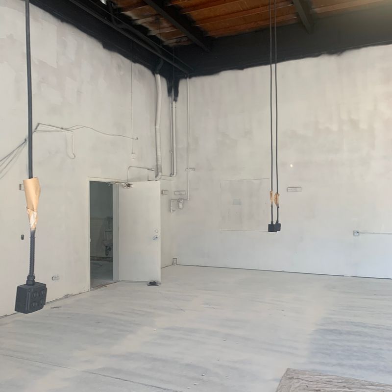 Empty industrial room with white walls, concrete floor, exposed black ceiling beams, and hanging cords.
