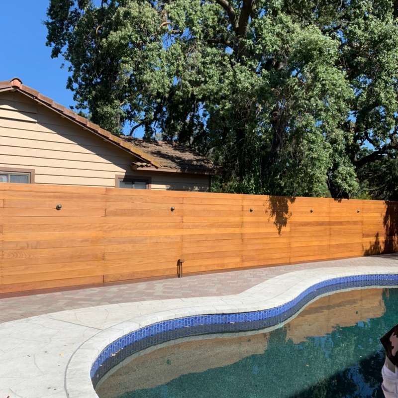 Backyard swimming pool with blue-tiled edge, concrete patio, wooden fence, and a house under trees.