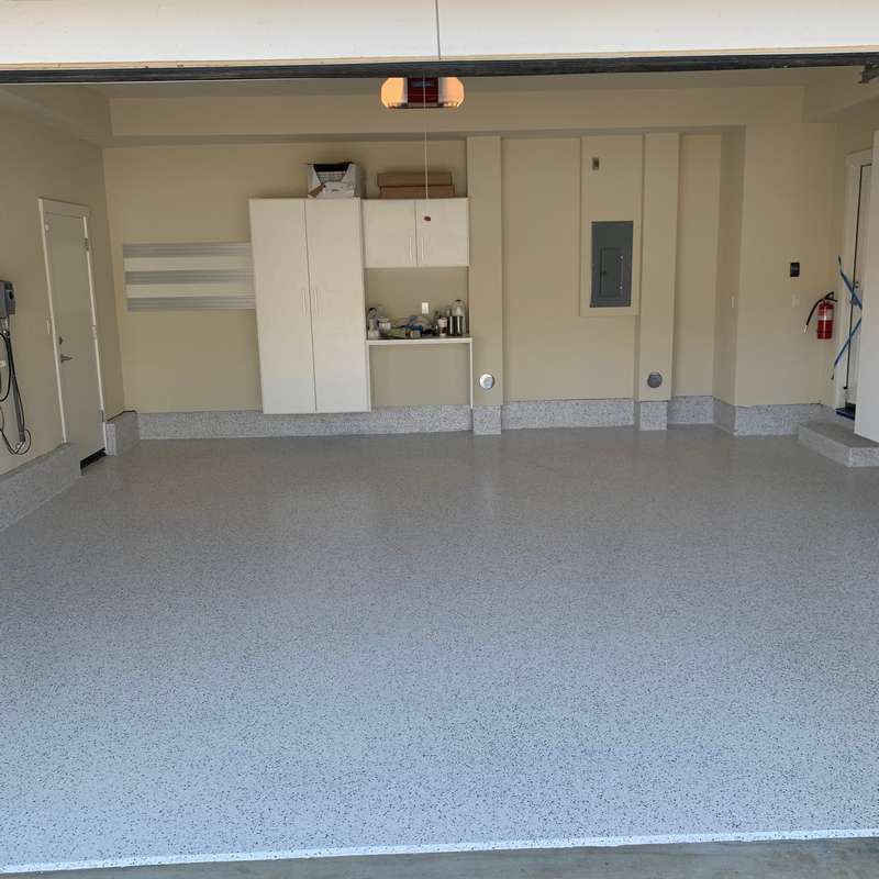Empty garage with gray speckled floor, white storage cabinets, and a side door on the back wall