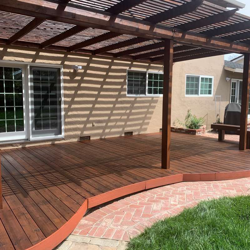 Backyard wooden deck with pergola, sliding glass doors, and curved brick edge beside green lawn