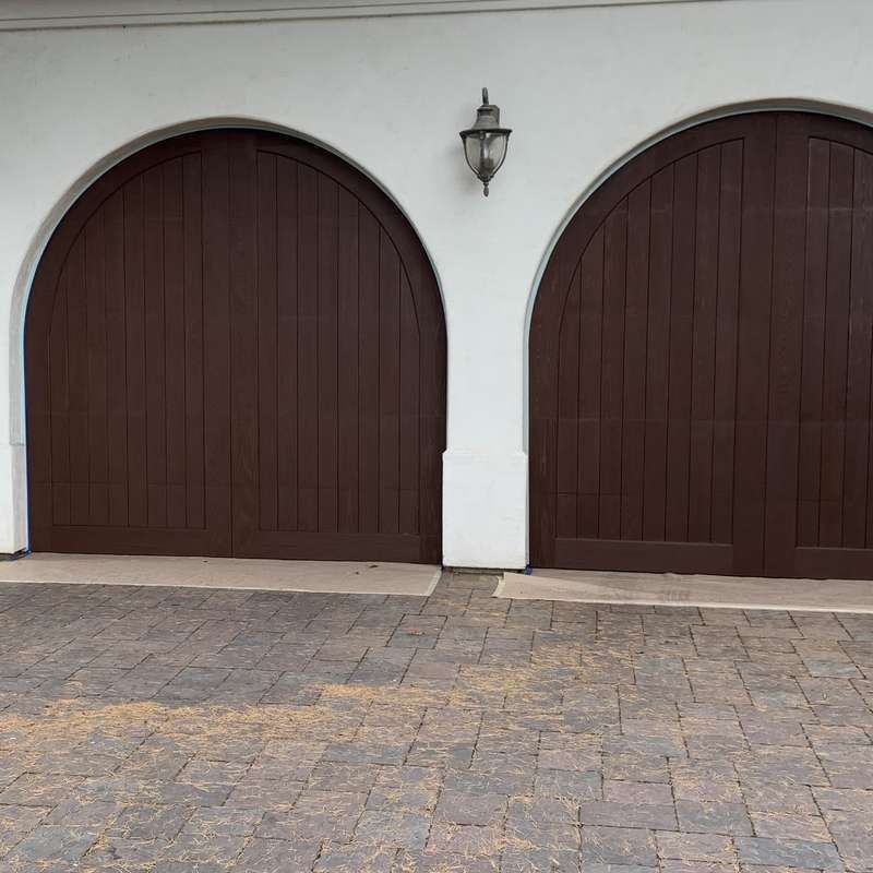 Two dark brown arched wooden doors set in a white stucco wall above a stone driveway