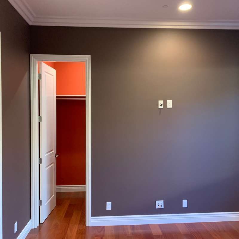 Empty room with gray walls, wood floor, white trim, and an open doorway to a red hallway.