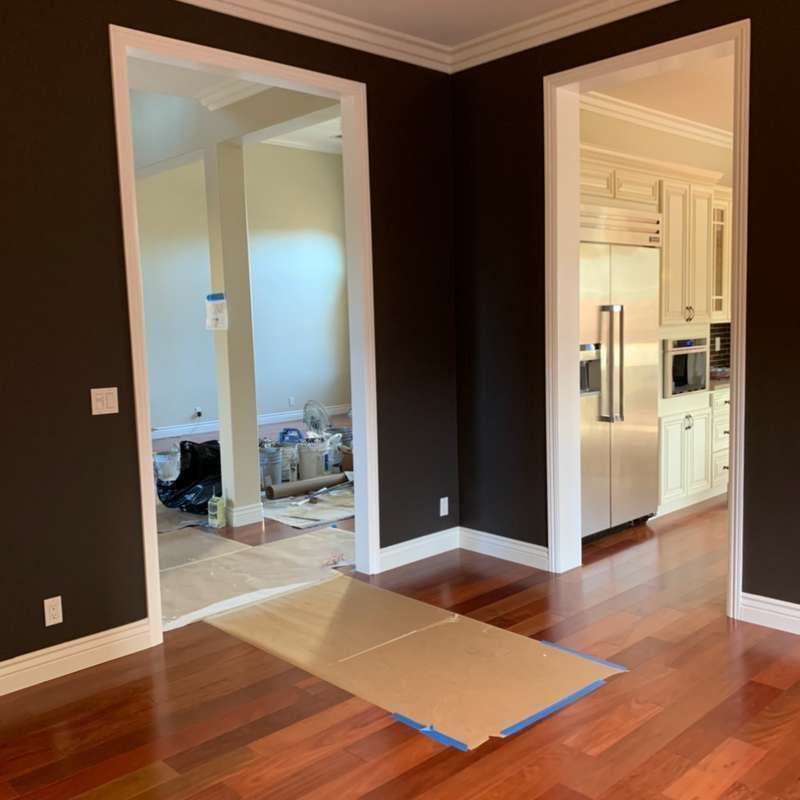 Empty room with dark walls, hardwood floors, and a cardboard sheet bridging two doorways into a kitchen.