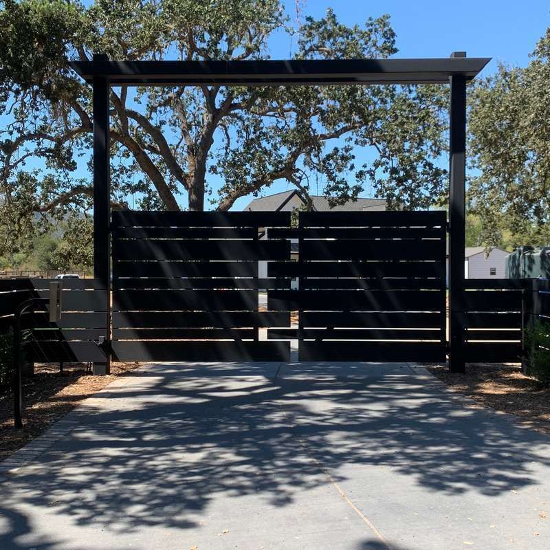Black wooden gate beneath a pergola, framed by trees and dappled sunlight on the ground.