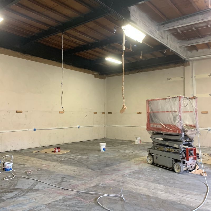 Empty warehouse under renovation with exposed ceiling, hanging cords, and construction equipment on the floor