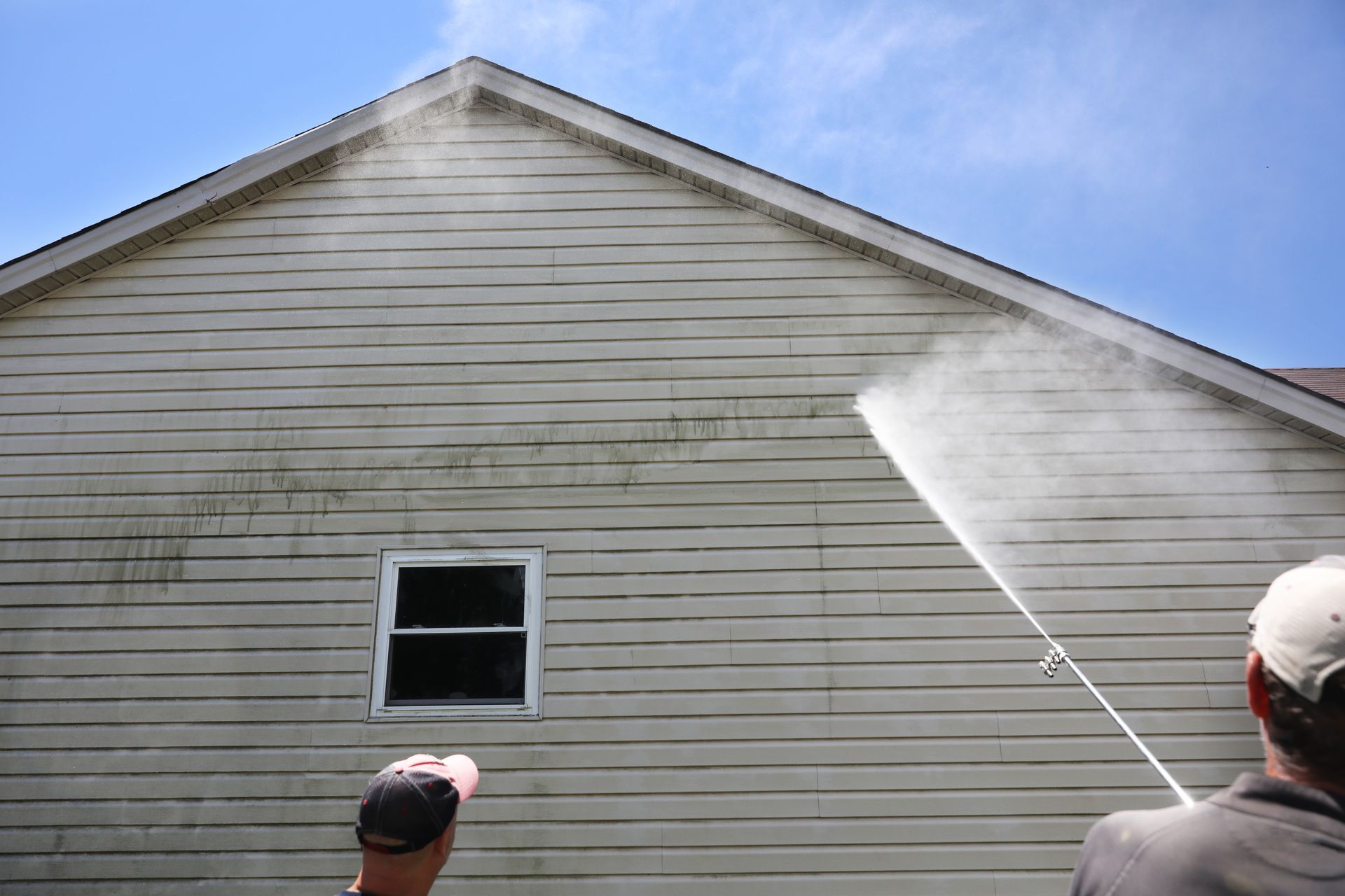 People power-washing the side of a beige house near a small window
