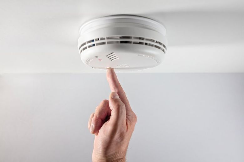 Hand pressing the test button on a white smoke detector mounted on a white ceiling.