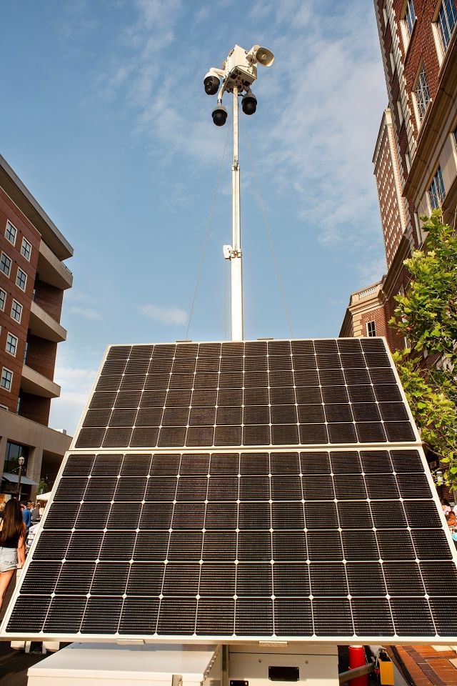 Solar-powered surveillance tower with cameras, between buildings, on a sunny day.