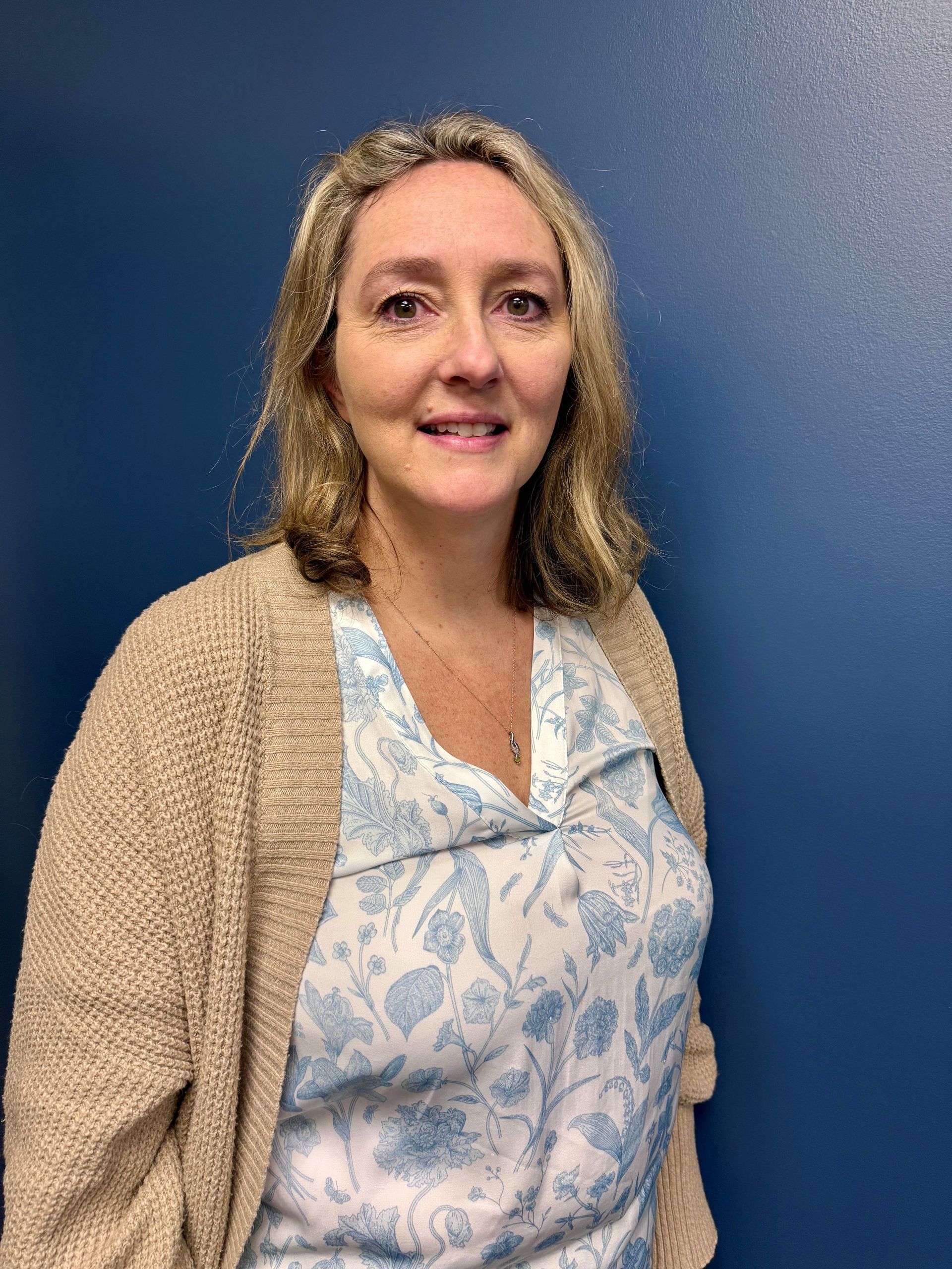 Woman in cardigan and floral shirt smiles in front of a blue wall.