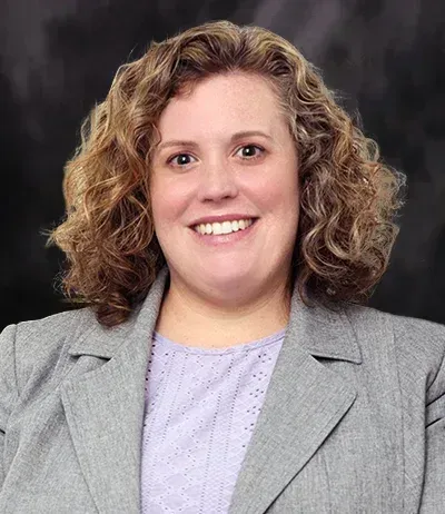 Woman with curly light brown hair smiles, wearing a gray blazer over a lavender top.