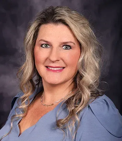 Woman with blonde wavy hair smiles, wearing a blue top, against a blurred background.