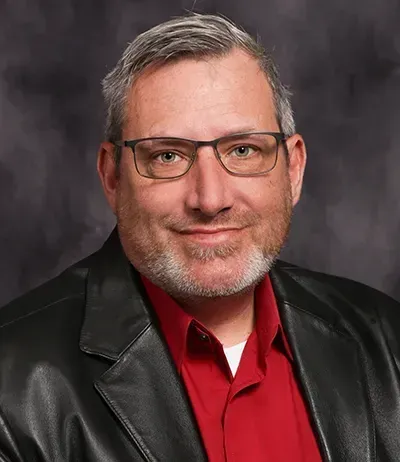 Man wearing glasses, red shirt, and black jacket, smiling in front of a blurred background.