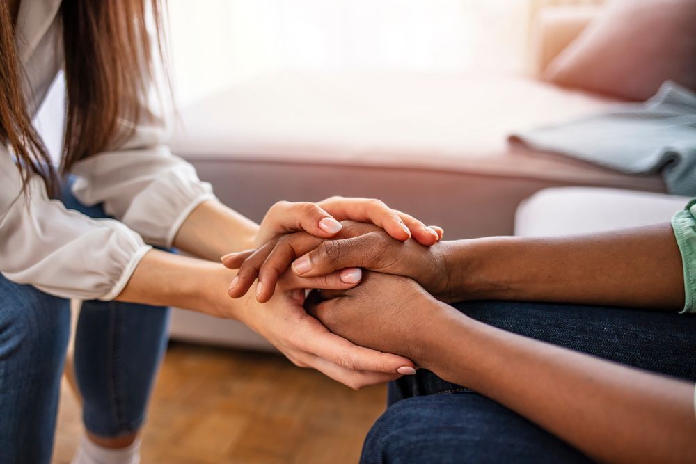 Two people's hands clasped together, possibly offering comfort. Indoors, near a sofa.