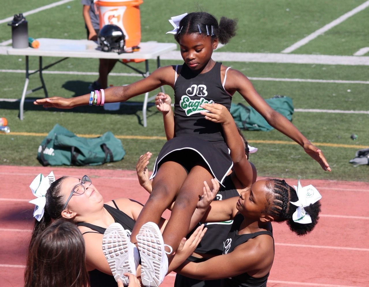 Flag football game at night, a player in red jersey runs with the ball as others pursue on a green field.