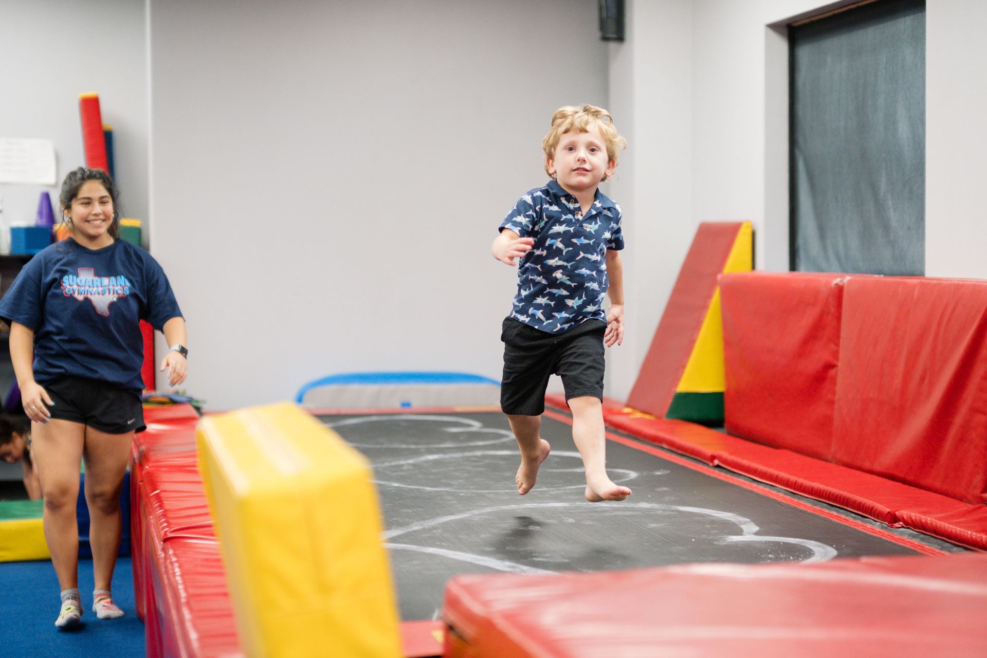 A young boy is jumping on a trampoline while a woman watches.