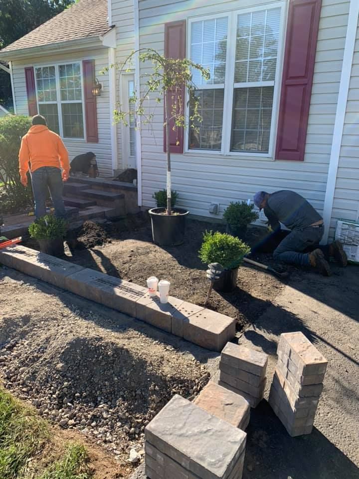A gardener wearing orange pants and gloves uses an electric hedge trimmer to shape a green hedge outdoors.