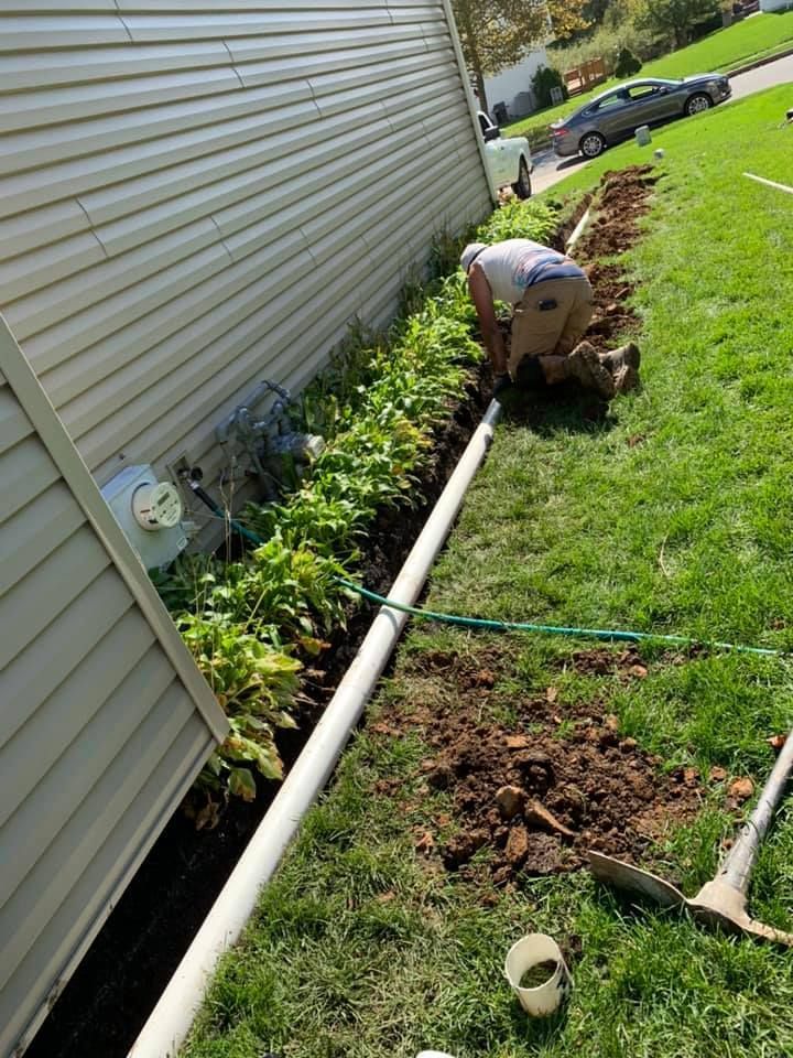 A person wearing green overalls and work gloves installs a roll of sod along a stone garden path in a backyard.