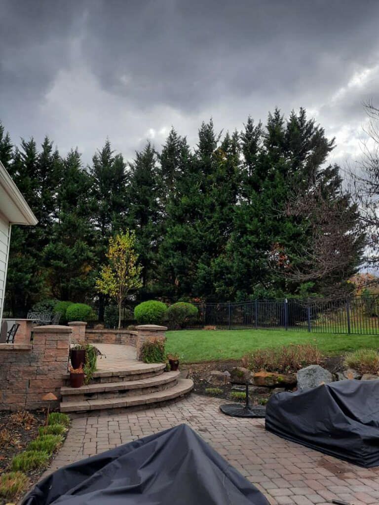 A stone pathway leads to the front door of a two-story home, flanked by lush green trees, shrubs, and colorful flowers.