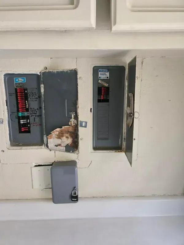 Electrical panels recessed in a wall; two metal panels, gray and black, with exposed wiring and damage around the edges.
