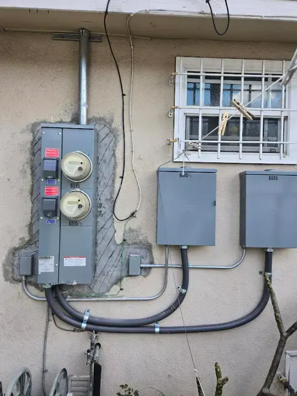 Electrical equipment on a wall with two meters, conduit, and metal boxes near a window with bars.