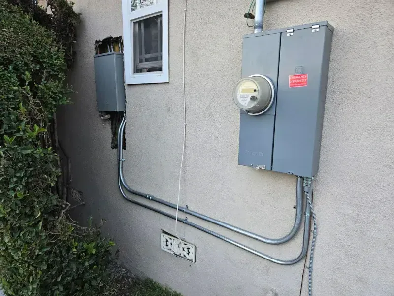 Electrical meters and boxes on a stucco wall beside a window and greenery.