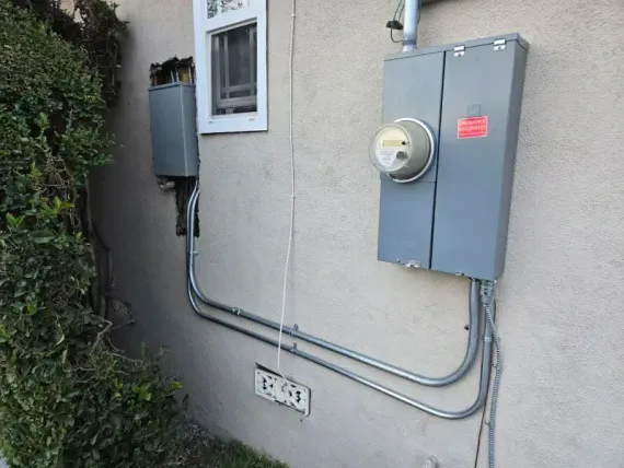 Electrical meters and boxes on a stucco wall beside a window and greenery.
