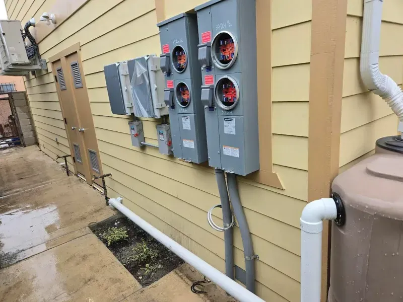 Electrical meters and equipment on a yellow building's exterior wall. A water tank and pipe are nearby.