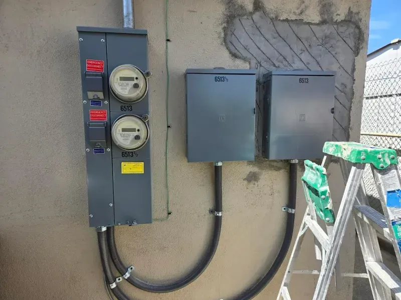 Electrical panel with two meters and two gray boxes on a stucco wall, ladder to the right.