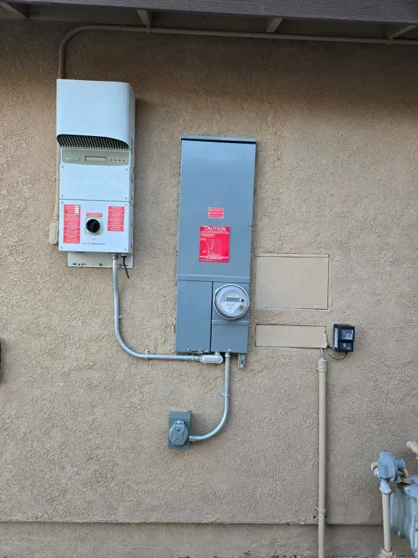 Gray electrical boxes and meter mounted on a beige stucco wall. Conduit pipes connect the boxes.