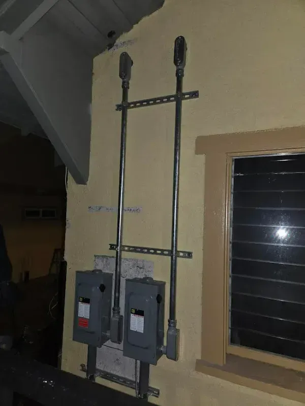 Two gray electrical boxes and conduits mounted on a yellow wall next to a window.