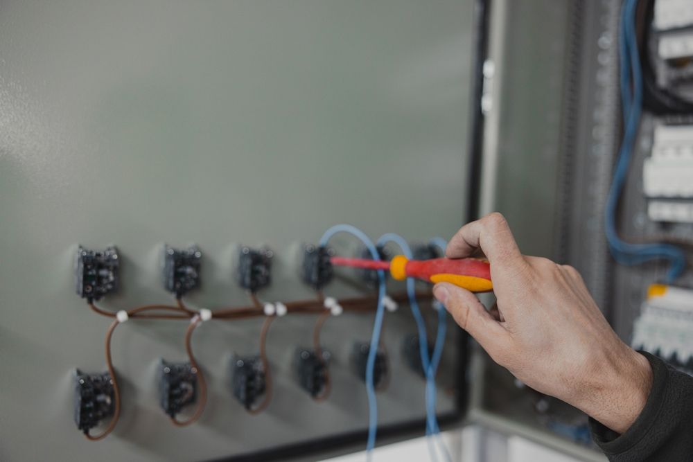 A person's hand using a screwdriver to work on electrical wiring inside a gray panel.