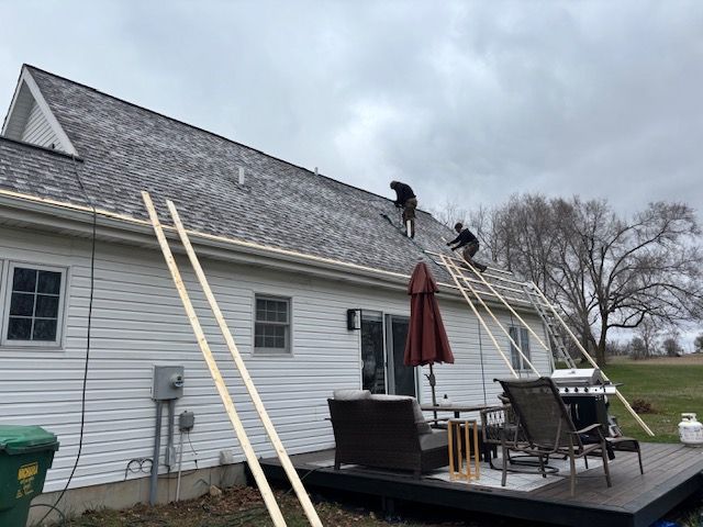 Two workers on a house roof with extension ladders leaning against the siding and a patio below