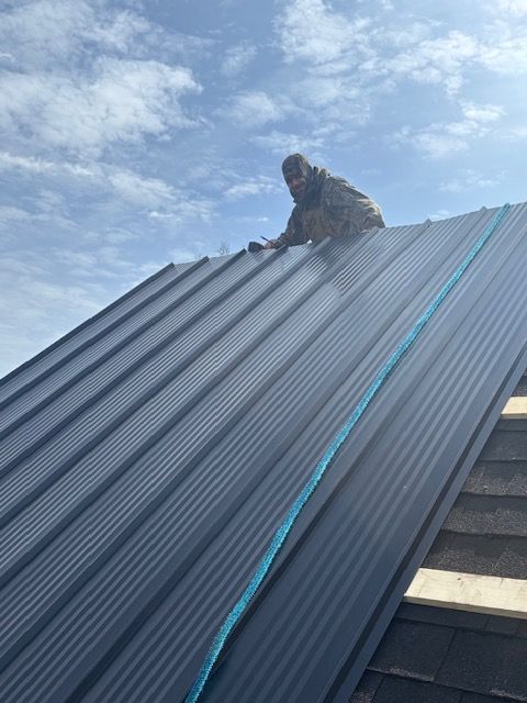 Worker installing dark metal roofing panels on a steep roof under a cloudy sky