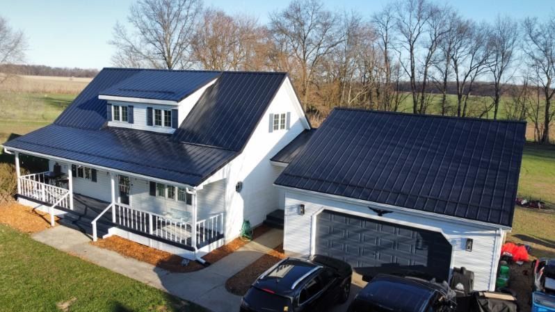 A two-story white house with a black metal roof, front porch, and attached garage in a rural, grassy setting.