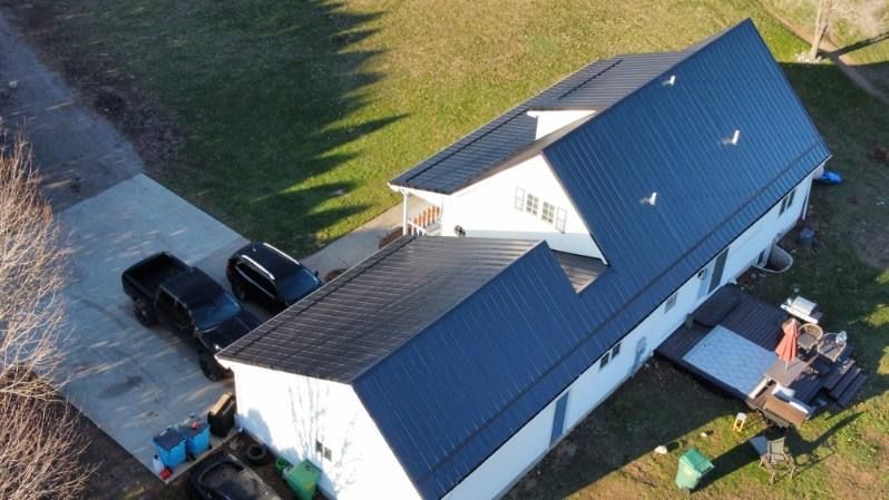 Aerial view of a white house with a dark metal roof, two parked trucks, and a small deck on a grassy lot.