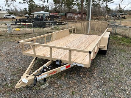A tan single-axle utility trailer with a new wooden deck and mesh rear gate parked on a gravel lot.