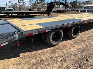 A flatbed trailer with wooden decking, metal side sections, and black tires parked on a gravel lot under a blue sky.