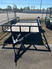 An empty utility trailer with a wooden floor and black metal frame sits on a gravel lot under a clear blue sky.