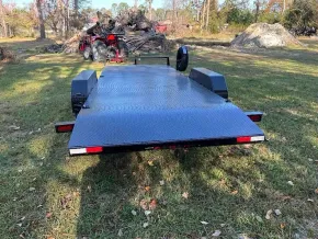 A black metal utility trailer with a diamond plate deck sits on a grassy field with a red vehicle parked in the distance.
