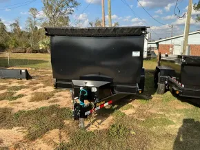 A black dump trailer parked on a grassy lot, viewed from the front, with a second trailer visible to the right.