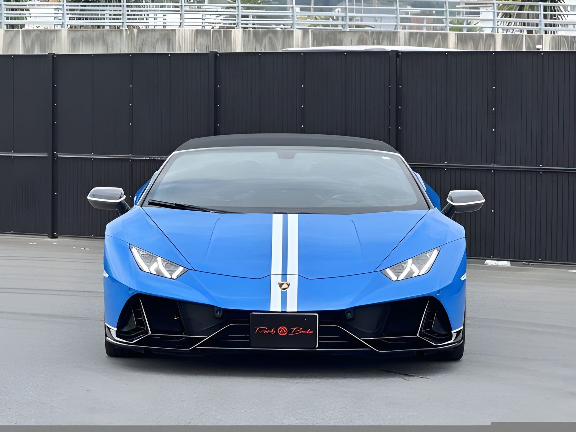 Blue Lamborghini sports car with white racing stripes; parked in front of a dark fence.
