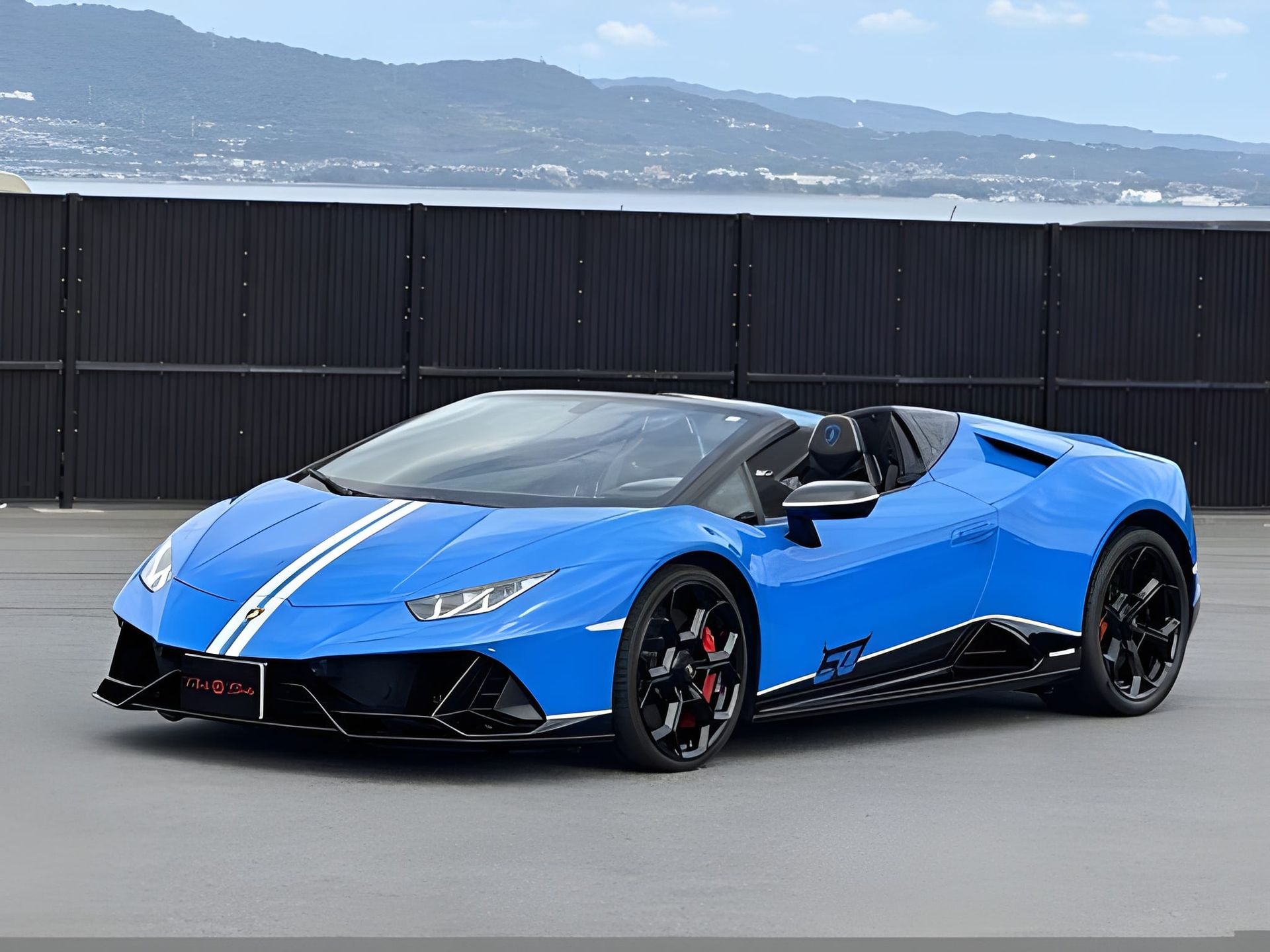 Blue Lamborghini sports car with white racing stripes; parked in front of a dark fence.