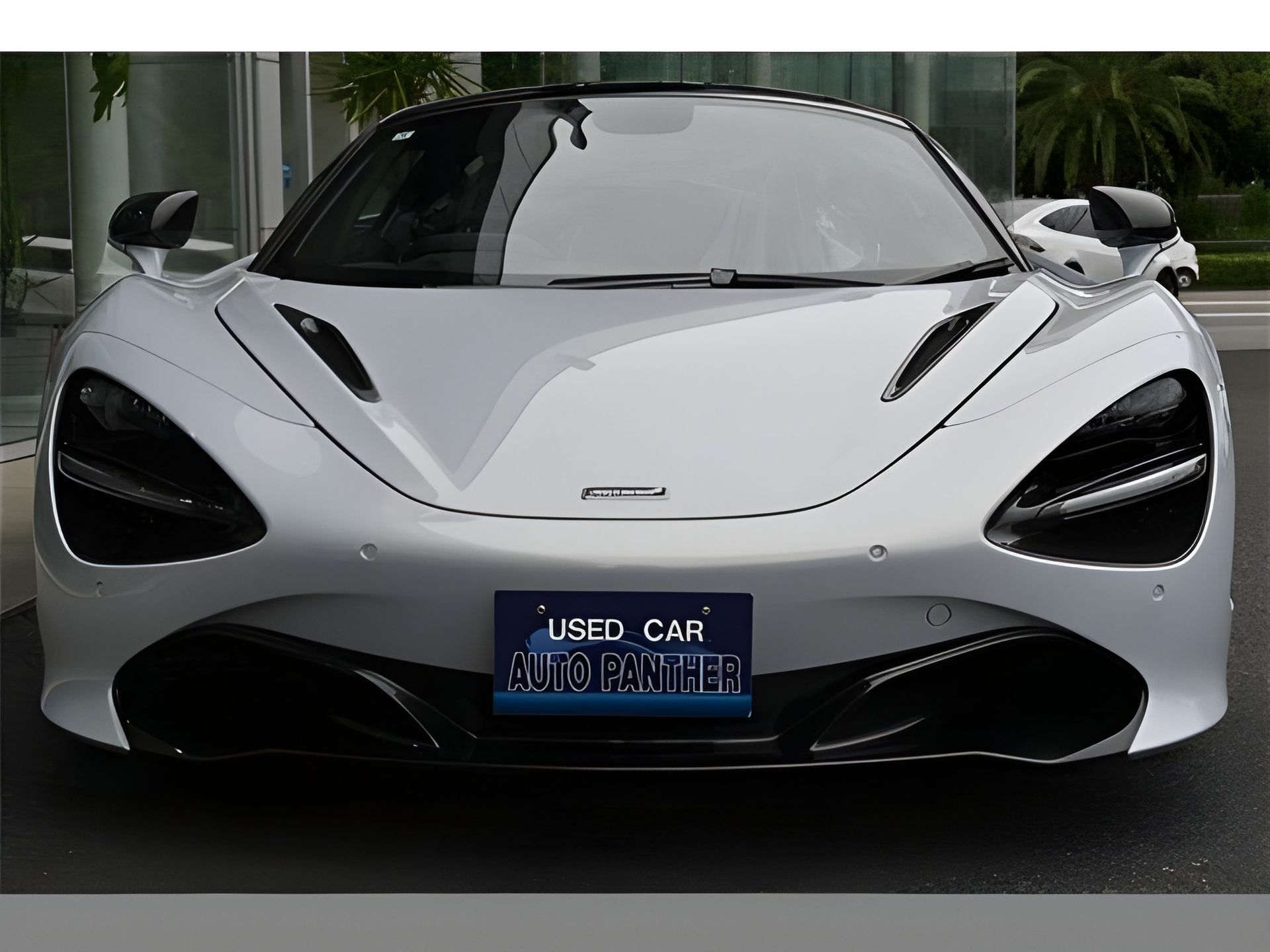 Silver McLaren sports car with black accents, parked in front of a building, displaying a "USED CAR" plate.