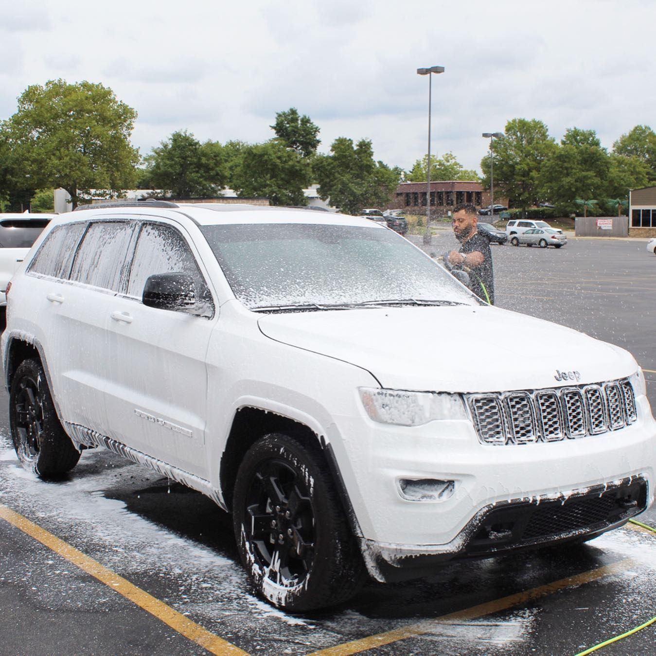 A white Jeep SUV covered in car wash foam parked in an outdoor lot, with a person nearby spraying water onto the vehicle.