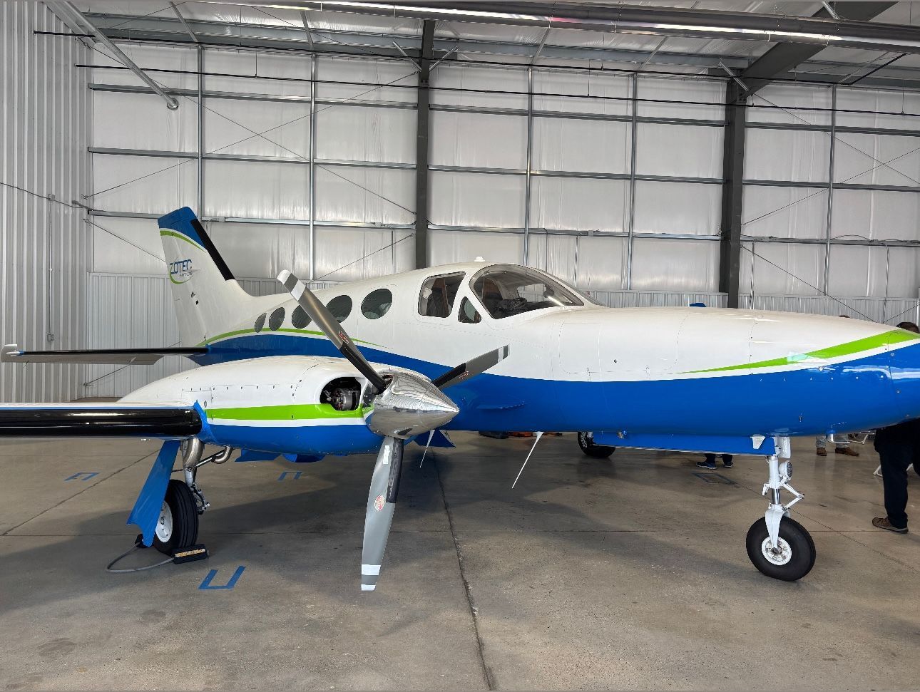A twin-engine propeller aircraft painted white, blue, and lime green parked inside a metal aircraft hangar.