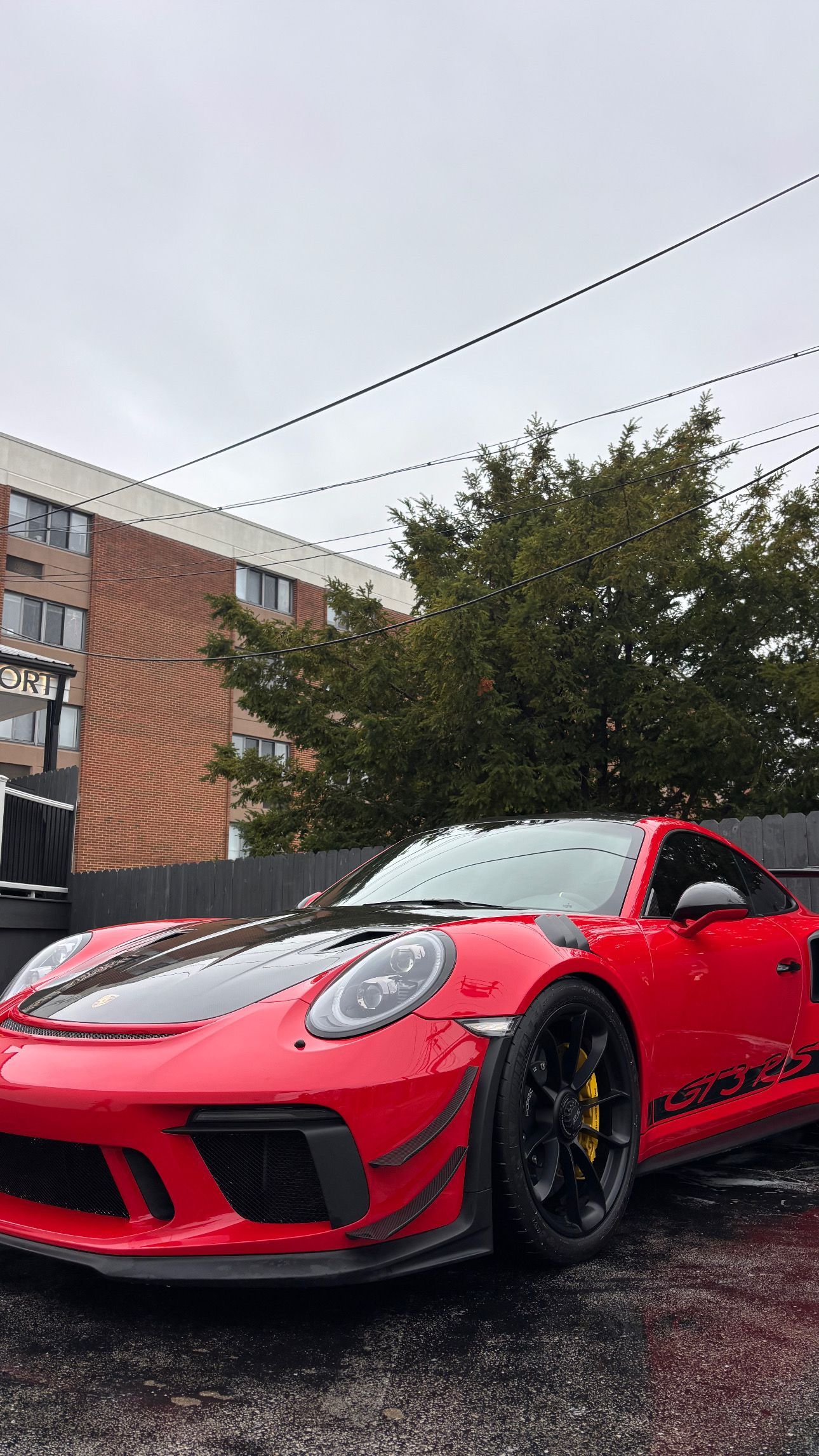 A red Porsche 911 GT3 RS parked outdoors on a cloudy day with a multi-story brick building in the background.