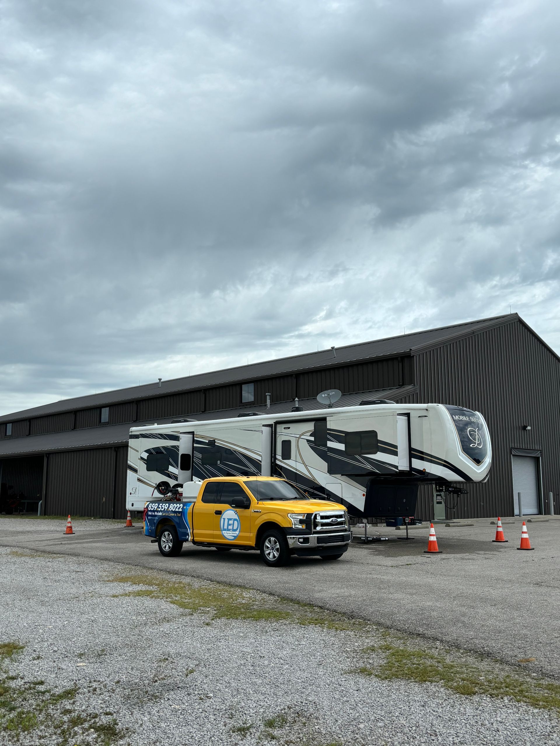 A yellow pickup truck hitched to a large white and black fifth-wheel RV parked on a gravel lot before a metal building.